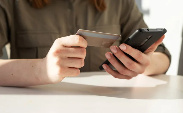 Ženský ruce u sebe držej kreditku a telefon. Žena sedí u stolu a platí v aplikaci, nakupování on-line nebo dělá bankovní transakce. — Stock fotografie