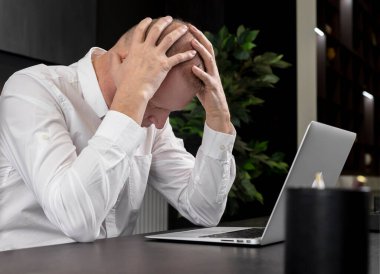 Ofis masasında dizüstü bilgisayarla oturan ve elleriyle yüzünü tutan yorgun bir işadamı. Çalışma başarısız, aşırı yükleme ve yanma kavramı