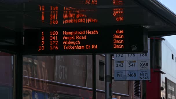 Een close-up van een digitaal bordje in een bushalte waarop staat welke bussen moeten rijden als Red London Double Decker Bussen op de achtergrond passeren — Stockvideo