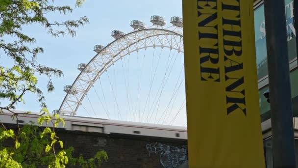 En vy ser upp på en järnvägsbro som ett tåg passerar med London Eye i bakgrunden och trädgrenar och en Southbank Centre skylt i förgrunden — Stockvideo
