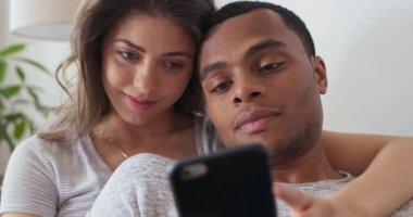 Kadın, erkek arkadaşına cep telefonunda bir şey gösteriyor. Kanepede dinlenirken. Afrika kökenli Amerikalı ve Kafkasyalı çift evde birlikte telefon kullanıyor. 4k ağır çekim el bilgisayarı