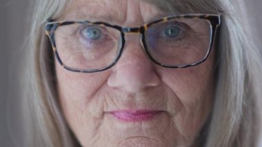 Gözlüklü, kameraya bakan ciddi derecede yaşlı, beyaz bir kadının portresi. ECU 'dan emekli yaşlı beyaz kadın pencerenin önünde. Yavaş çekim 4k