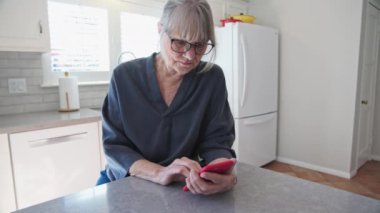 Üst düzey beyaz kadın mutfak tezgahında otururken cep telefonuyla mesaj ya da e-posta atıyor. Evde akıllı telefonuyla çalışan yaşlı beyaz kadın. Yavaş çekim 4k