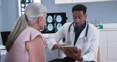 Genç Afro-Amerikalı doktor, kafa yaralanması belirtileri hakkında not almak için tablet kullanıyor. Kafkasyalı kadın hastanın beyin sarsıntısı hakkında genç bir doktora danışması.