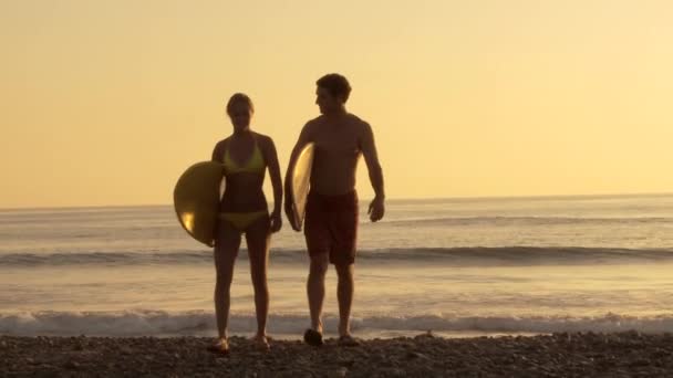 夕暮れ時にビーチにサーフボードを持つ若いカップル — ストック動画