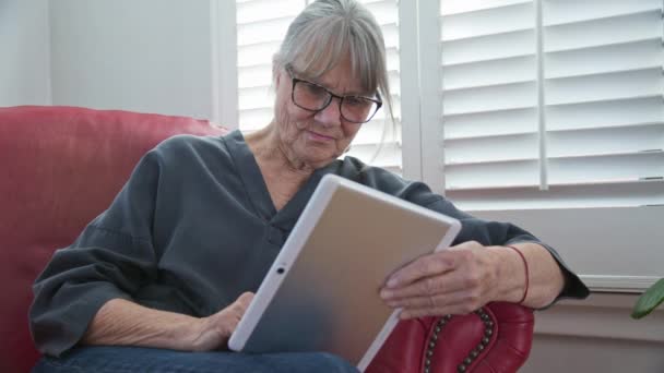 リビングルームに座っている間 タブレットコンピュータを使用してシニア白人女性 モバイルデバイスでアプリを使用してニュースをチェックしたり インターネットを閲覧する高齢の白人女性 スローモーション4K — ストック動画