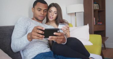 Genç çift, kredi kartı ve akıllı telefonla online alışveriş yapıyor. Milenyum Afrikalı Amerikalı ve Kafkasyalı yeni evliler tatil rezervasyonu yapıyor. 4k ağır çekim el bilgisayarı