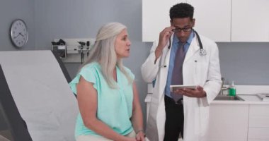 Yaşlı kadın hasta tıp odasında oturuyor ve genç siyahi doktora danışıyor. Milenyum erkek doktoru, üst düzey bir hastayla konuşurken elinde elektronik tablet tutuyor.