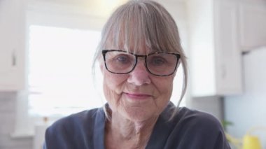 Dost canlısı, beyaz saçlı, gözlüklü, beyaz saçlı ve mutfakta kameraya bakan son sınıf beyaz kadın portresi. Yaşlı, beyaz bir bayanın evinde başından ve omzundan vuruldu. Yavaş çekim el bilgisayarı 4k