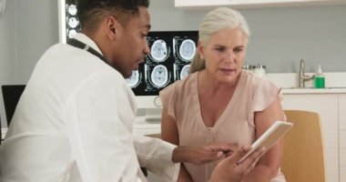 Tıp doktoru ve son sınıf öğrencisi, kapalı bir sağlık kliniğinde tablet bilgisayara bakıyor. Genç siyah doktor bilgisayarlı tomografiden kadın hasta test sonuçlarını göstermek için elektronik ped kullanıyor.