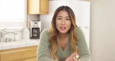 Asyalı kadın mutfakta otururken kameraya bakıp sohbet ediyor.