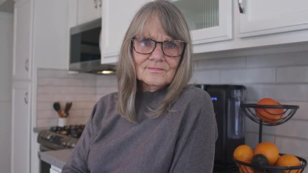 中等近距离的画像灰色头发的老年妇女与眼镜看着相机在她的厨房 身穿灰色毛衣的年长的白人退休人员 慢动作4K — 图库视频影像