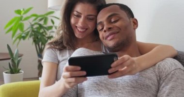 Cep telefonuyla video izlerken kanepede kıvrılıp yatan sevimli genç bir çift. Afrika kökenli Amerikalı ve Kafkasyalı çift birlikte cep telefonu kullanıyor. 4k ağır çekim el bilgisayarı