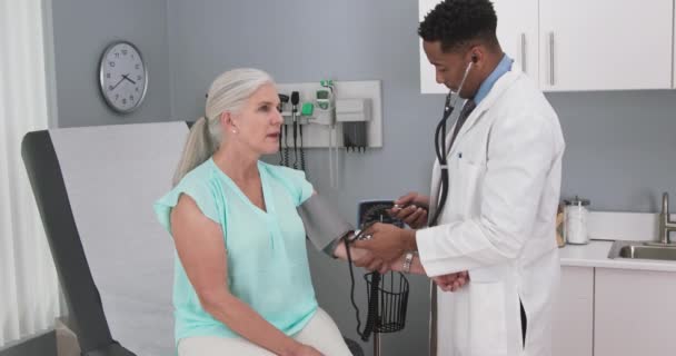 老年高加索病人的血压由千禧年的非洲裔美国医生检查 年轻男性医生使用监视器和听诊器测量老年妇女的血压 — 图库视频影像