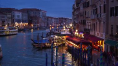 Şehir ışıklarının bulanık arkaplanı ve akşamları Büyük Kanal 'da yürüyen insanlar. Kanal boyunca kemerli pencereleri ve balkonları olan renkli Venedik binaları. 4k