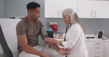 Tıp odasında oturan yakışıklı Afro-Amerikalı hasta kan basıncını kıdemli doktora kontrol ettiriyor. Yetişkin beyaz kadın doktor, erkek hastaların kan basıncını kontrol ediyor.