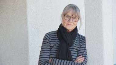 Kendine güvenen kıdemli ev sahibi evinin önünde duruyor bir sütuna yaslanıyor ve kameraya bakıyor. Yaşlı, beyaz, gözlüklü, çizgili süveter ve eşarplı bir kadın. Yavaş çekim 4k