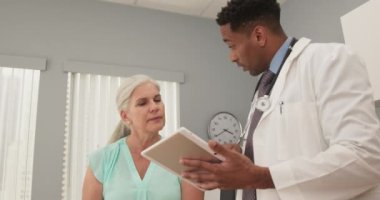 Genç Afro-Amerikalı doktor, beyaz kadın hastaya danışmanlık yaparken tablet bilgisayar kullanıyor. Milenyum doktoru elektronik ped kullanıyor ve yaşlı bir kadınla konuşuyor.
