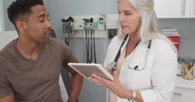 Hasta notları almak için tablet kullanan olgun beyaz kadın doktor. Genç erkek Afrikalı-Amerikalı hasta, kapalı bir sağlık kliniğinde kıdemli doktorla görüşüyor. El bilgisayarı ağır çekim