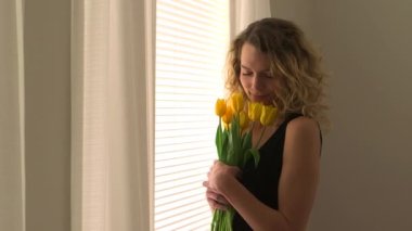 Bir buket çiçekle pencerenin yanında duran sarışın kadın.