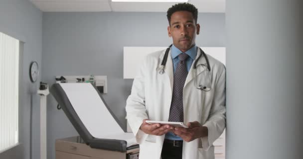 英俊的非洲裔美国男医生的画像靠在诊所的墙上 漂亮的黑人医生在医生办公室拿着平板电脑 看着相机 — 图库视频影像