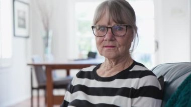 Evde kameraya bakan, ciddi derecede yaşlı, beyaz bir kadını yakından çek. Oturma odasında oturan çizgili kazaklı, gri saçlı son sınıf öğrencisi. Yavaş çekim 4k