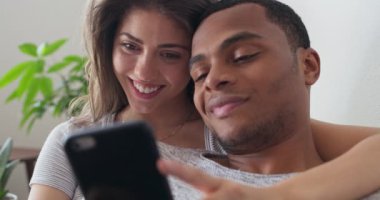 Genç milenyum çifti, kanepede dinlenirken kız arkadaşlarının telefonundan şirin bir video izliyor. Afro-Amerikalı ve beyaz kadın akıllı telefonu birlikte kullanıyorlar. 4k ağır çekim el bilgisayarı