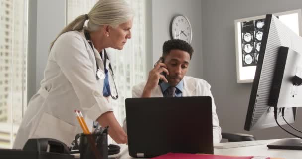 若いアフリカ系アメリカ人医師が医療室内の携帯電話を使っている 患者の医療ファイルに若い同僚と働くシニア女性医師 — ストック動画