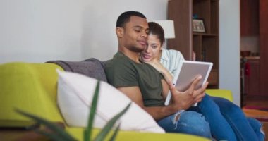 Mutlu genç milenyum çifti koltukta tablet bilgisayara bakıp gülüyor. Çekici Afrikalı Amerikalı ve Kafkasyalı çift cep telefonu kullanıyor ve gülümsüyor. 4k ağır çekim el bilgisayarı