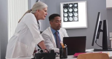 Milenyum Afrikalı-Amerikalı doktor kıdemli Kafkasyalı meslektaşıyla notlarını karşılaştırıyor. Bilgisayarları kullanarak hastaların tomografilerini paylaşan iki doktor.