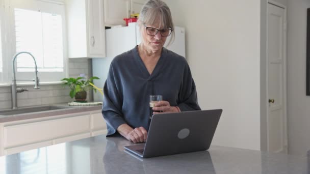 アイスコーヒーを飲みながらキッチンでノートパソコンを使って笑顔の年上の白人女性のドリーショット カウンターでパソコンで働く先輩白人女性の中出し スローモーション4K — ストック動画