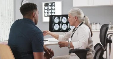 Genç Afro-Amerikalı hasta, beyin sarsıntısı hakkında kıdemli tıp doktoruyla görüşüyor. Yaşlı kadın doktor hastanın beyin tomografisini gösteriyor.