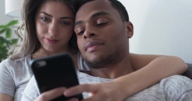 Kız arkadaşı, erkek arkadaşına cep telefonunda bir şey gösteriyor. Adam kanepede ona yaslanıyor. Afrika kökenli Amerikalı ve Kafkasyalı çift evde birlikte telefon kullanıyor. 4k ağır çekim el bilgisayarı