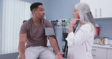 Çekici erkek Afro-Amerikan hasta kapalı bir klinikte kan basıncına baktırıyor. Olgun kadın doktor, genç erkek hastanın kan basıncını ölçüyor.
