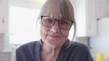 Mutfakta kameraya gülümseyen beyaz saçlı, gözlüklü, yaşlı beyaz bir kadının portresi. Mutlu, yaşlı, emekli bir bayanın evinde başından ve omzundan vurulduğu fotoğraf. Yavaş çekim el bilgisayarı 4k