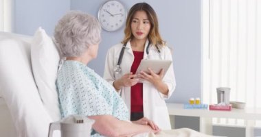 Asyalı doktor elinde tablet bilgisayarla hastane yatağında olgun bir kadınla konuşuyor.