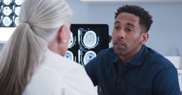 高齢の医師に耳を傾け男性患者の閉じると 脳震盪の治療を説明します 脳損傷のX線について若い男性患者と話す高齢女性医師の肖像 — ストック動画