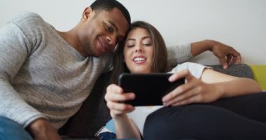 Sosyal medyada telefonla sevimli resimlere gülen genç bir milenyum çifti. Afrikalı Amerikalı ve Kafkasyalı sevgili internetten komik videolar izliyor. 4k Yavaş çekim el bilgisayarı