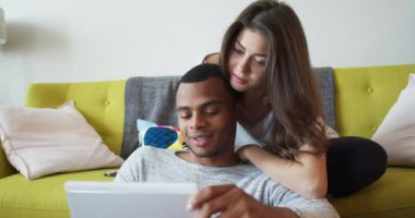 Genç milenyum çifti oturma odasındaki koltukta birlikte tablet bilgisayar kullanıyorlar. Çevrimiçi cihaz kullanan Afrikalı Amerikalı ve Kafkasyalı sevgili. 4k Yavaş çekim el bilgisayarı