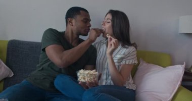 Çekici genç Afrikalı Amerikalı ve beyaz çift kanepede birbirlerini patlamış mısırla besliyorlar. Milenyum erkeği ve kadın oturma odasında birlikte atıştırmalık yiyor. 4k ağır çekim el bilgisayarı