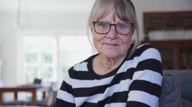 Evde kameraya bakan kendinden emin, sağlıklı, yaşlı, beyaz bir kadın var. Oturma odasında oturan çizgili kazaklı, gri saçlı son sınıf öğrencisi. Yavaş çekim 4k