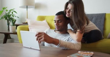 Mutlu genç milenyum çifti oturma odasında tablet bilgisayar kullanıyor. Afro-Amerikalı ve Kafkasyalı erkek arkadaş ve kız arkadaş apartmanda video izliyorlar. 4k Yavaş Hareket El Bilgisayarı