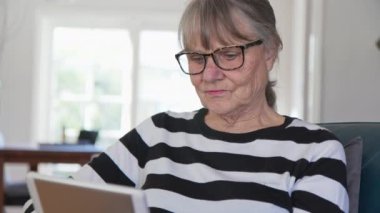 Oturma odasında otururken tablet bilgisayar kullanan mutlu Kafkasyalı kadın. Yaşlı beyaz kadın e-okuyucudaki uygulamayı haberleri kontrol etmek ya da internette gezinmek için kullanıyor. Yavaş çekim 4k