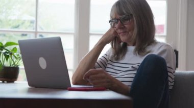 Kafkasyalı mutlu bir kadın yemek masasında dizüstü bilgisayarla çalışırken rahat rahat oturuyor. Yaşlı beyaz kadın mesajlaşırken ya da sevdiklerine e-posta gönderirken gülümsüyor. Yavaş çekim 4k