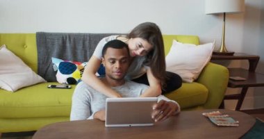 Erkek arkadaşı, nişanlısını oturma odasında tutarken tablet bilgisayarında bir şey gösteriyor. Afrika kökenli Amerikalı ve Kafkasyalı Milenyum çifti mobil cihazları paylaşıyor. 4k ağır çekim el bilgisayarı