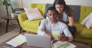 Makbuzları gözden geçirip bütçelerini gözden geçiren genç bir çift. Afrika kökenli Amerikalı ve Kafkasyalı sevgililer oturma odalarında faturalarını internetten ödüyorlar. 4k Yavaş çekim el bilgisayarı