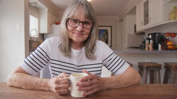 キッチンテーブルで朝のコーヒーを片手にカメラを見ている幸せなシニア白人女性の肖像画 優しい引退した女性は自宅でお茶のマグカップを楽しんでいます スローモーションドリーショット4K — ストック動画