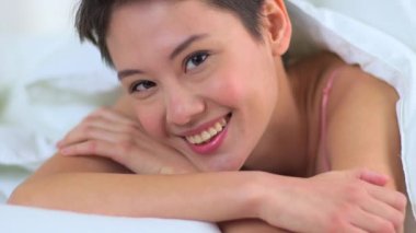 Yatakta çarşafların altında gülümseyen melez Asyalı kadın.