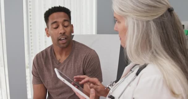 魅力的な黒人男性患者は首の痛みで医師の訪問を支払う 患者の健康のメモを取るためにポータブルタブレットを使用して高齢者の白人医師の閉鎖 — ストック動画