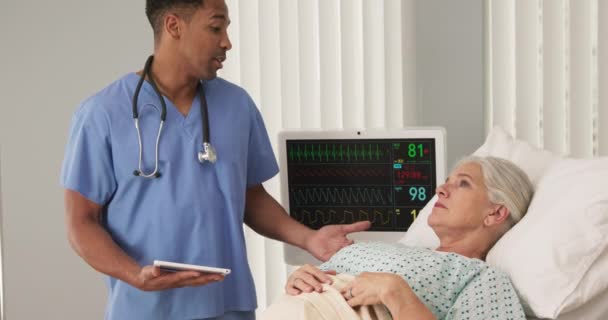 老年病患者卧床期间男性黑人护士使用平板电脑的肖像 躺在医院病床上的老年妇女 与年轻护士交谈时与心电图监视器相连 — 图库视频影像